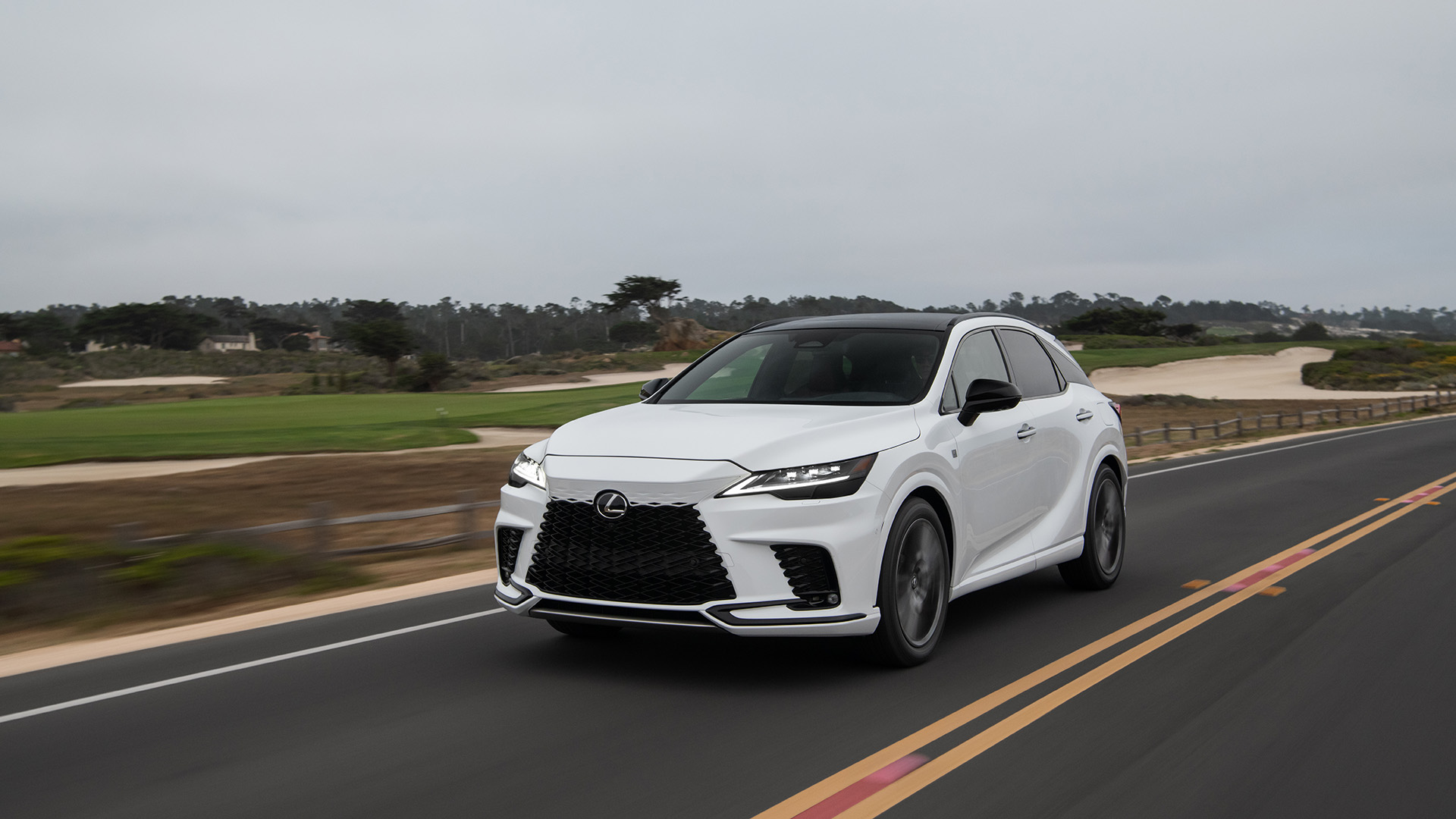 A white 2023 Lexus RX drives down the road near a golf course.